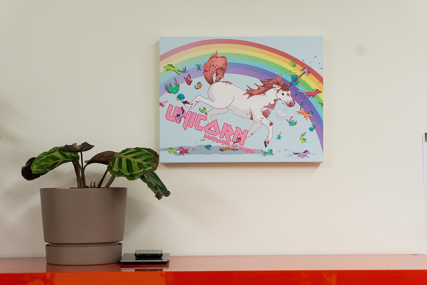 A simple plant and a couple of devices sit in front of a unicorn artpiece reading 'Unicorn: Destroyer of Ponies'